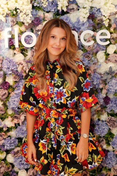 Millie Bobby Brown stands in front of a colorful floral background wearing a vibrant patterned dress, HD Mobile Wallpaper