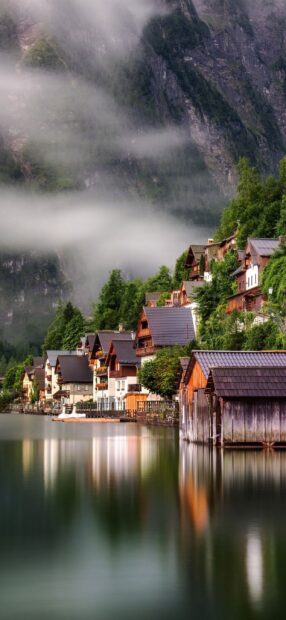 A mist covered village with wooden houses reflects on a calm lake surrounded by mountains in Austria, 2K Phone Wallpaper
