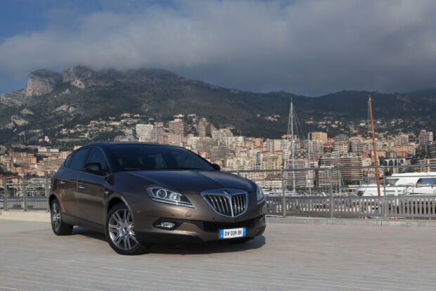 A sleek Lancia car is parked by a marina with a city and mountains in the background, 2K Desktop Wallpaper