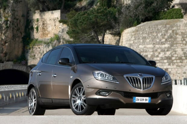 A brown modern Lancia car is parked near a stone wall and trees, 2K Desktop Wallpaper