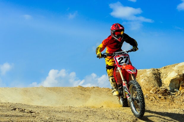 A motocross rider wearing red and yellow gear speeds on a dirt track under a blue sky, HD Desktop Wallpaper