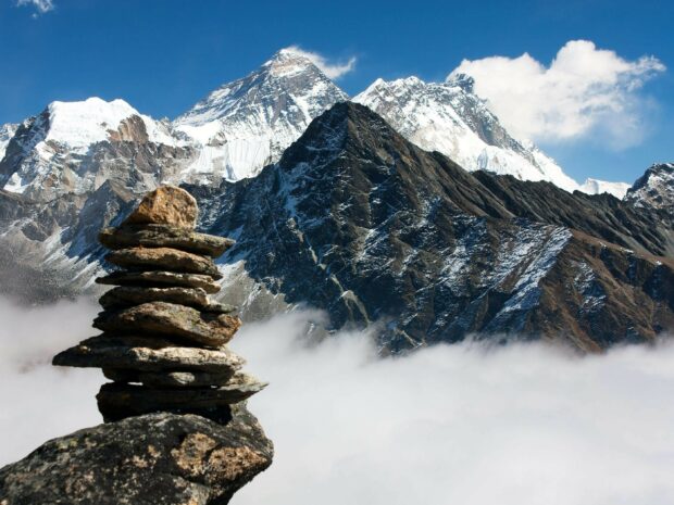 A stone cairn stacked on a rock with Mount Everest towering in the background above the clouds
