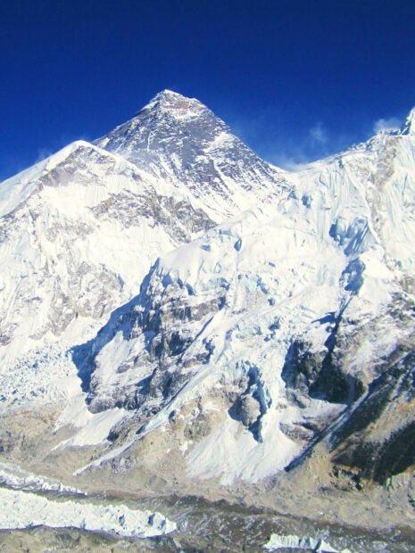 Snow covered Mount Everest peak under clear blue sky with rugged terrain visible