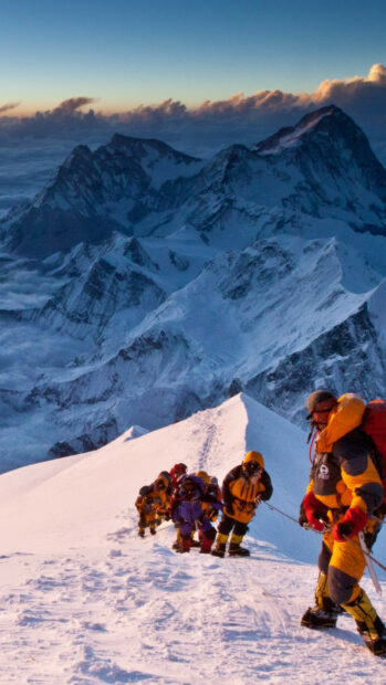 Climbers ascending a snowy Mount Everest peak in bright gear against a backdrop of rugged peaks and clouds
