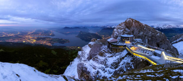 A snowy view of Mount Pilatus with buildings and a city illuminated in the distance at dusk