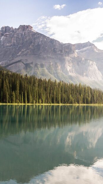 A mountain lake reflects tall pine trees and rocky cliffs under a partly cloudy sky, 2K Mobile Wallpaper
