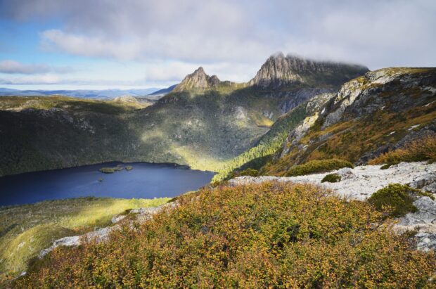A mountain lake rests below rocky hills with autumn foliage under a cloudy sky, 2K Desktop Wallpaper