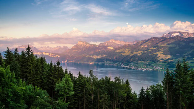 A peaceful mountain lake scene with dense evergreen trees and distant peaks under a cloudy sky, 4K Desktop Wallpaper