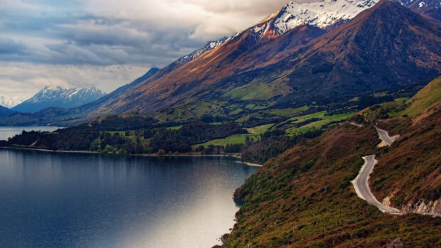 A mountain lake reflects the surrounding hills and snow capped peaks under a cloudy sky, 4K Desktop Wallpaper