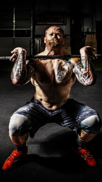 A muscular man with tattoos is performing a heavy powerlifting squat in a gym, 2K Phone Wallpaper