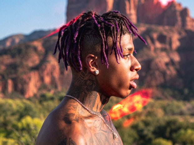 A side profile of Nle Choppa with purple dreadlocks and tattoos in a natural outdoor setting