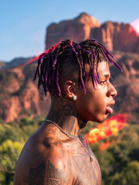 Side profile of Nle Choppa with purple dreadlocks and tattoos in a desert landscape background