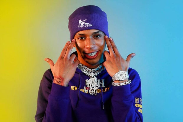 Nle Choppa posing with hand gesture wearing purple beanie and chains on a colorful background