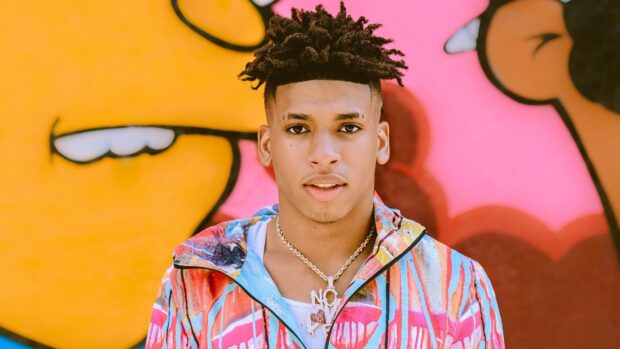 Nle Choppa standing in front of a colorful graffiti wall wearing a vibrant jacket