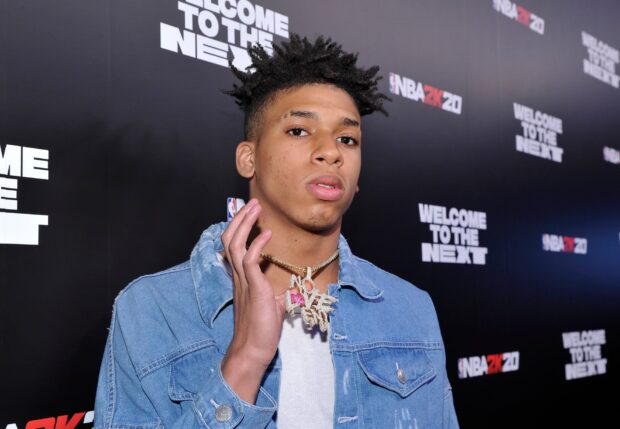 Nle Choppa wearing a denim jacket and a gold necklace in front of a promotional backdrop