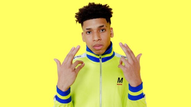 Nle Choppa wearing a yellow and blue jacket posing with both hands against a yellow background