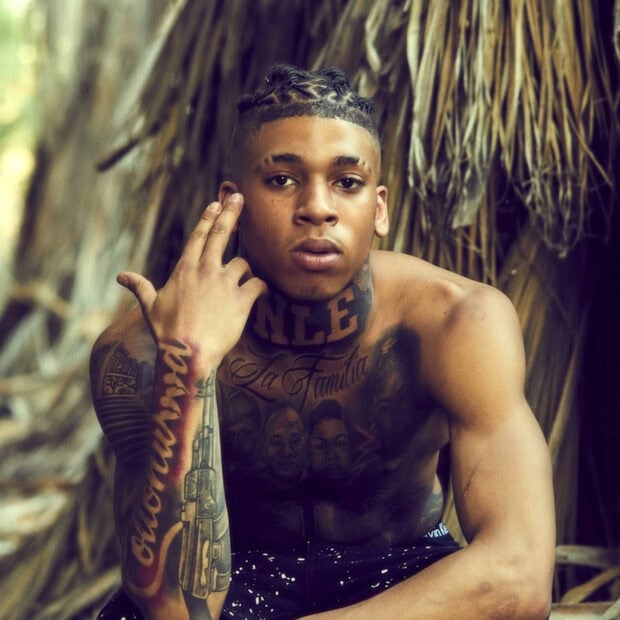 Nle Choppa sitting shirtless with detailed tattoos in an outdoor natural setting