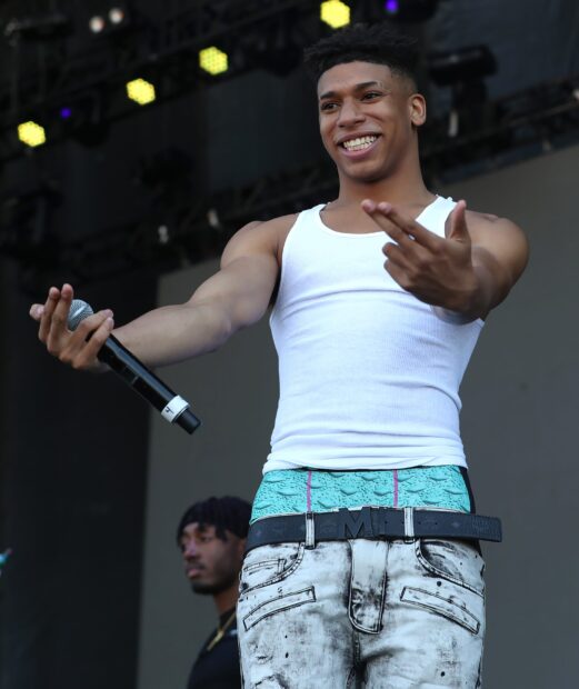 Nle Choppa smiling and holding a microphone on stage wearing a white tank top