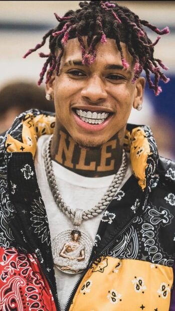 A smiling man with nle tattoo on his neck and unique hairstyle wearing jewelry and patterned jacket