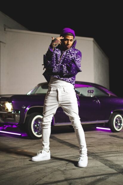 Nle Choppa wearing a purple beanie and jacket standing in front of a classic car