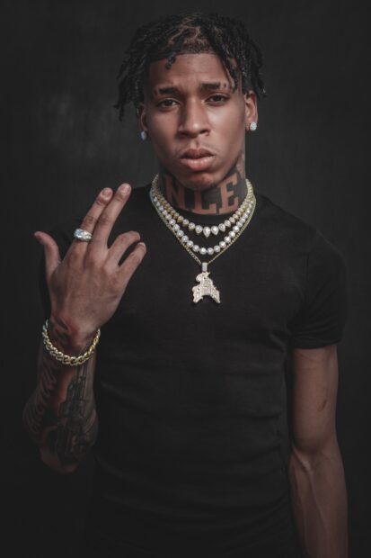 Nle Choppa wearing multiple diamond necklaces and a black shirt with tattoos on his arm and neck showing a hand gesture