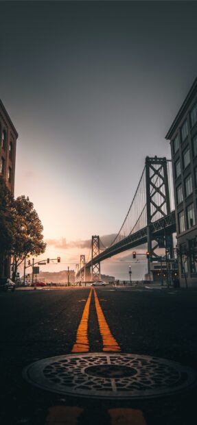 The Oakland bridge towers above a street with yellow road lines at sunset