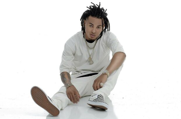 A man wearing casual white clothes and checkered shoes sits on the floor, showing tattoos and jewelry in this Ozuna Wallpapers