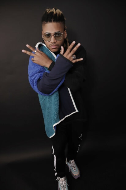 A man wearing sunglasses and colorful jacket poses confidently against a dark background with hands crossed