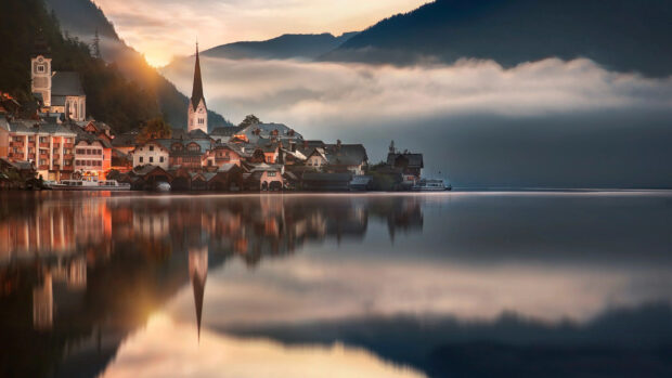 A peaceful Austrian village with a church tower reflects on the calm lake surface surrounded by mountains, 4K Desktop Wallpaper