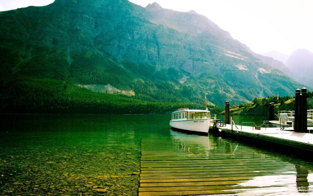 A boat is docked by the pier with mountains and clear lake waters in Glacier National Park, 2K Desktop Wallpaper
