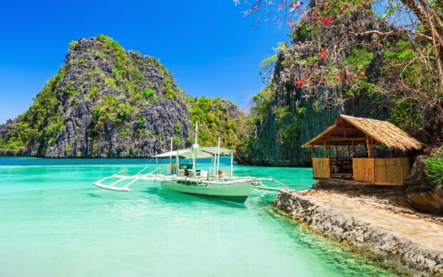 Traditional Philippine boat floating near tropical limestone cliffs and bamboo hut
