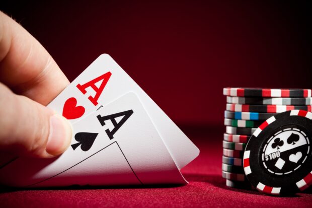 A hand holding a pair of aces in poker beside a stack of poker chips on a red table