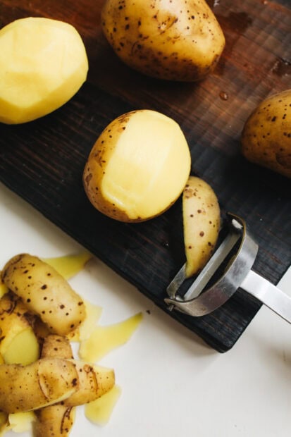 A peeled potato on a dark wooden board next to a peeler and potato peels on a white surface