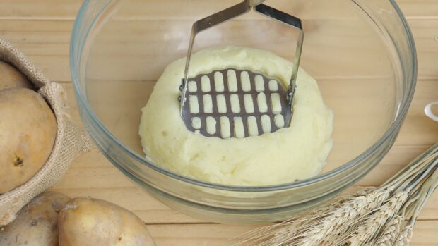 Mashed potato being prepared with a potato masher in a glass bowl on a wooden surface