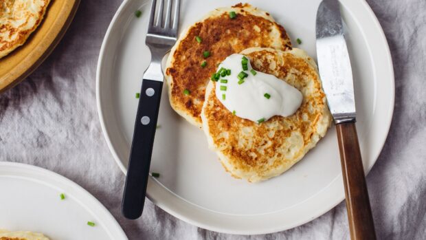 Two potato pancakes topped with sour cream and chopped chives on a white plate with fork and knife