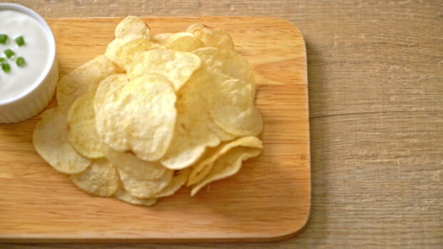 A stack of crispy potato chips served on a wooden board next to a bowl of dip with green onions
