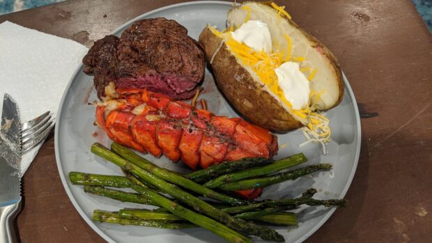 A baked potato topped with sour cream and cheese on a plate with lobster tail steak and asparagus