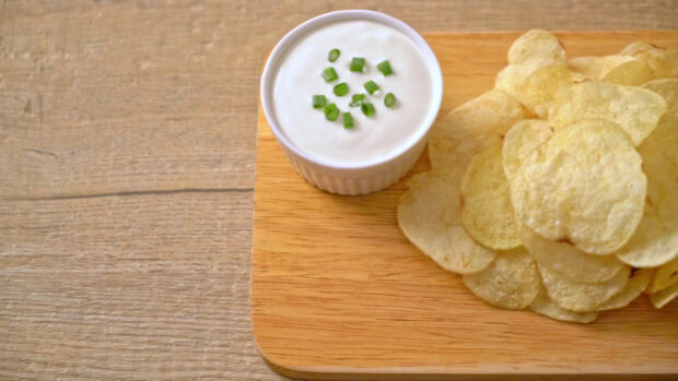 A bowl of creamy dip with chopped green onions served with crispy potato chips on a wooden board