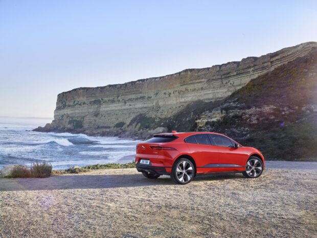 A red Jaguar I Pace is parked by the ocean with cliffs in the background during clear daylight, 2K Desktop Wallpaper