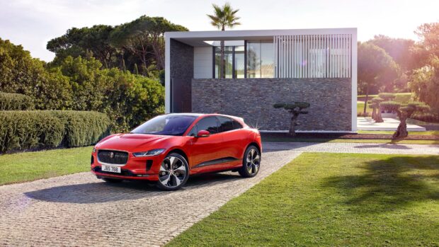 A red Jaguar I Pace car is parked on a driveway in front of a modern house surrounded by greenery, 4K Desktop Wallpaper
