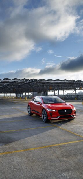 A red Jaguar I Pace is parked in an empty rooftop parking lot under a partly cloudy sky, 2K Phone Wallpaper