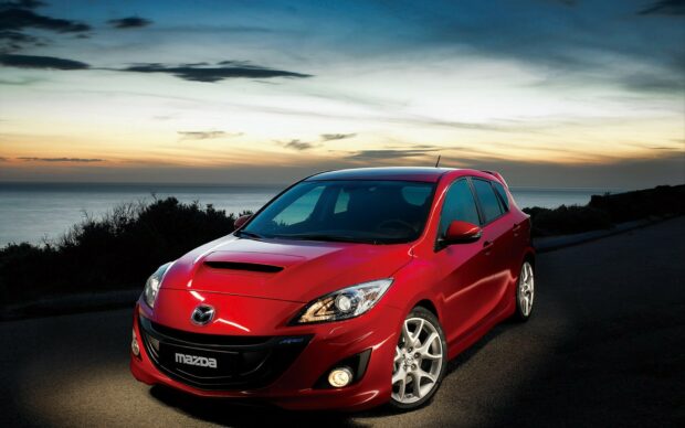 A red Mazda hatchback is parked on a coastal road with the ocean and sunset sky in the background, HD Desktop Wallpaper