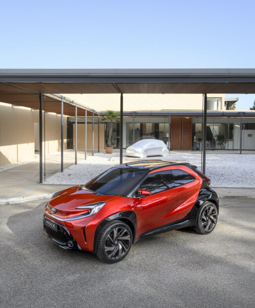 Futuristic red and black compact electric car parked in front of modern house with sleek architectural design.