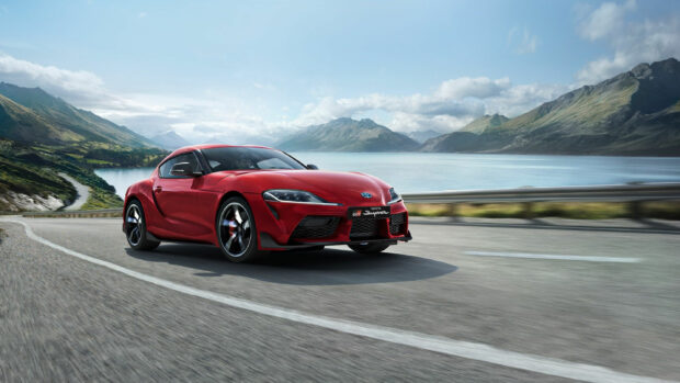 Red Toyota GR Supra sports car on an open road near a lake with mountains in the background.