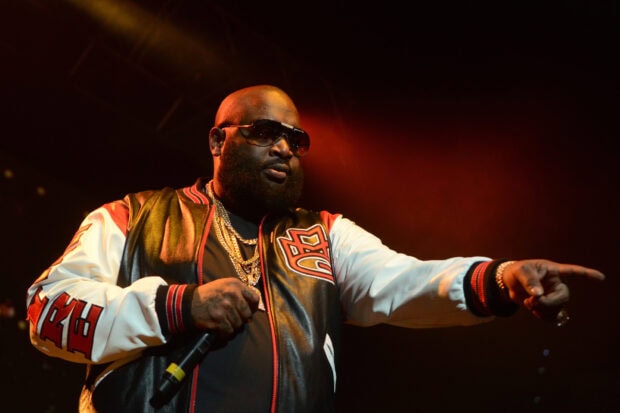 Rick Ross is performing on stage while pointing with his hand in front of a dark background, HD Desktop Wallpaper