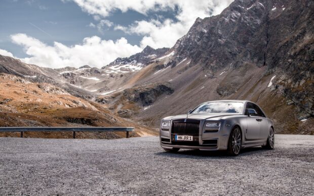 A Rolls Royce Ghost is parked on a mountain road with rocky peaks in the background, 2K Desktop Wallpaper