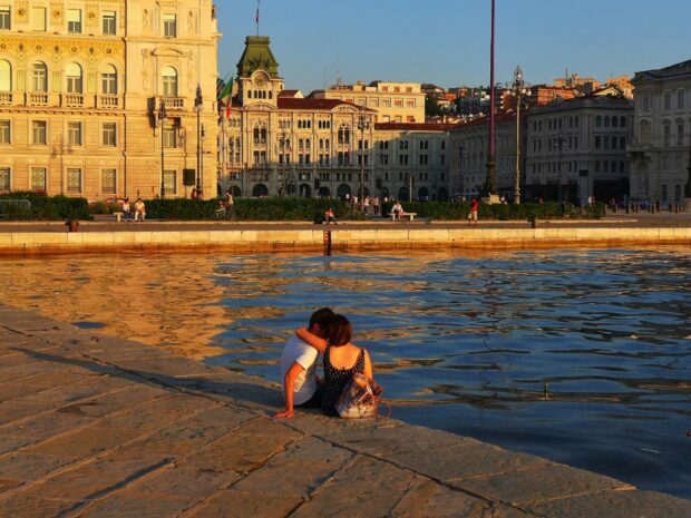 A couple enjoys the sunset by the waterfront in Trieste with historic buildings in the background, HD Desktop Wallpaper
