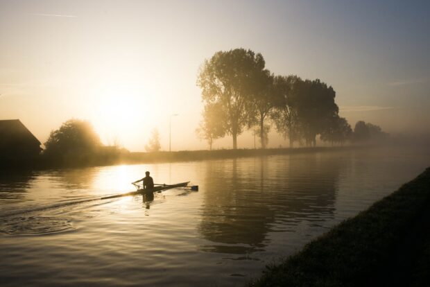 A rowing athlete paddles calmly on a misty river at sunrise with trees lining the shore
