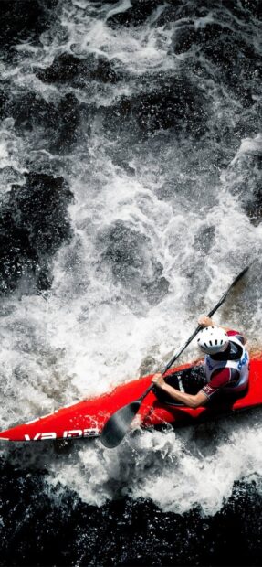 A kayaker in a white helmet paddles through turbulent water in a bright red kayak