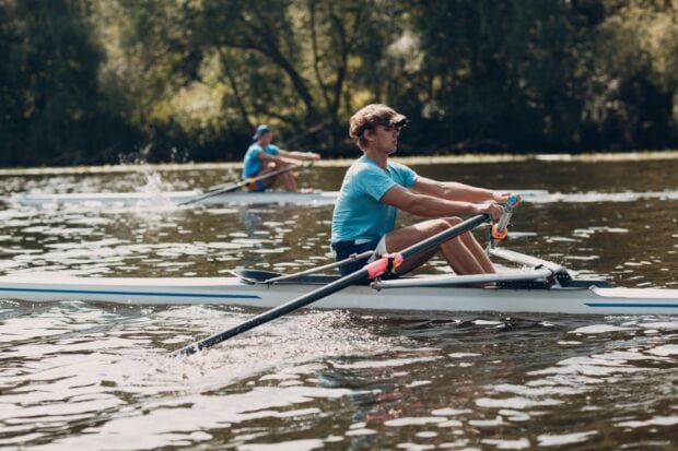 A young man rowing on a calm river with another rower in the background